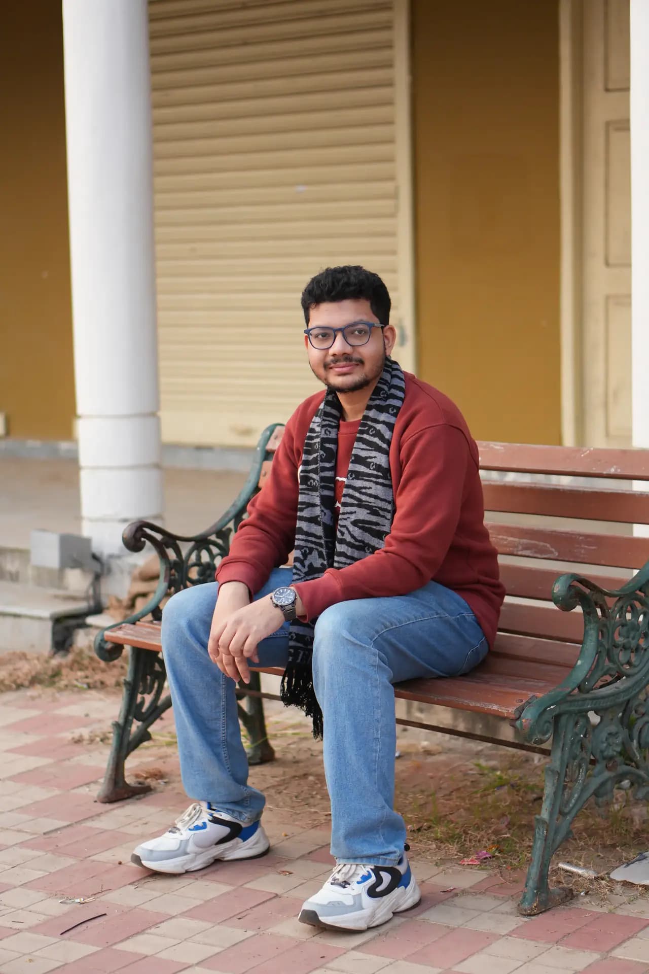 Raktim Ranjit sitting on a park bench in front of a yellow shuttered door, wearing a red sweater and scarf