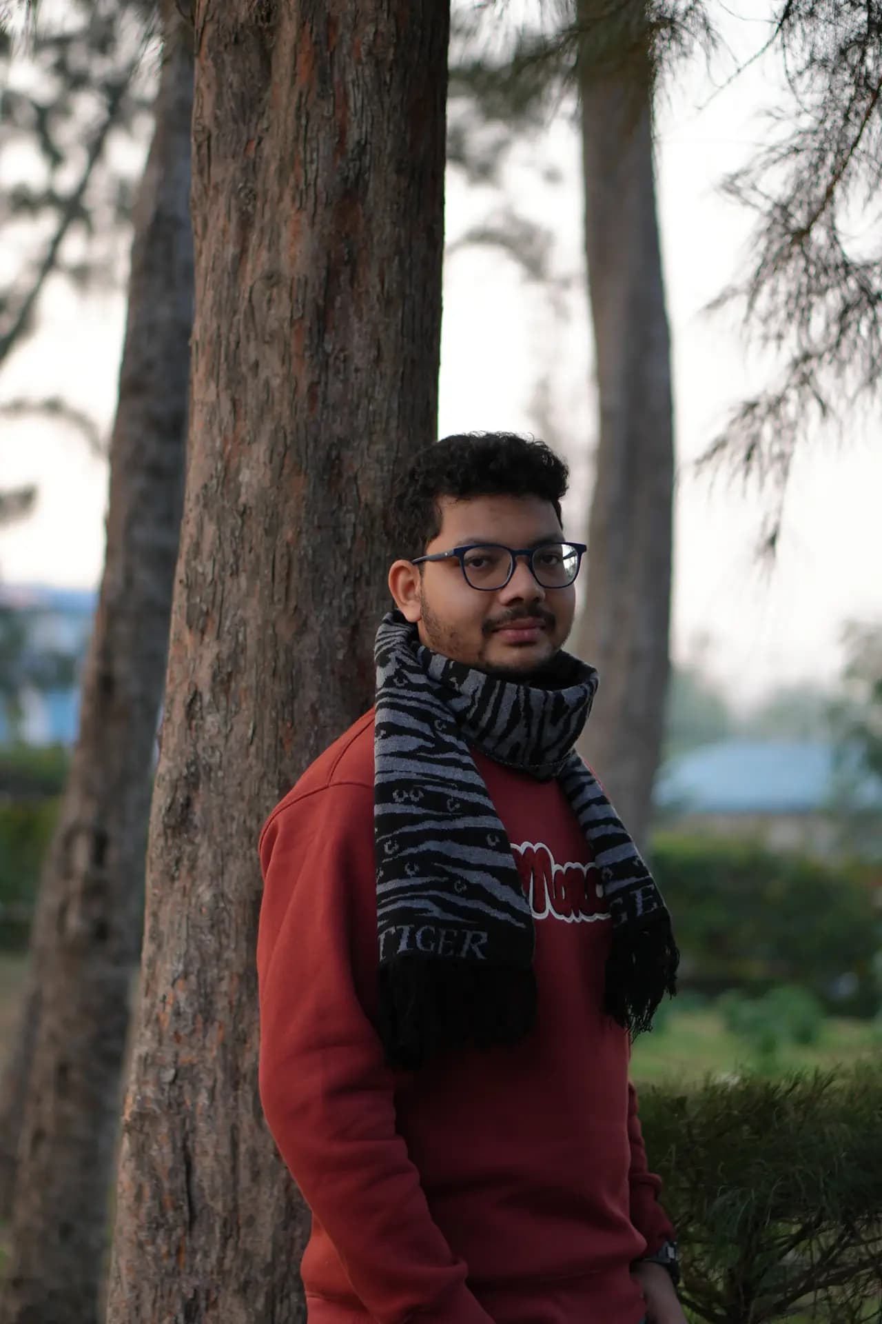 Raktim Ranjit in a red sweater and patterned scarf, half hidden behind a large tree trunk