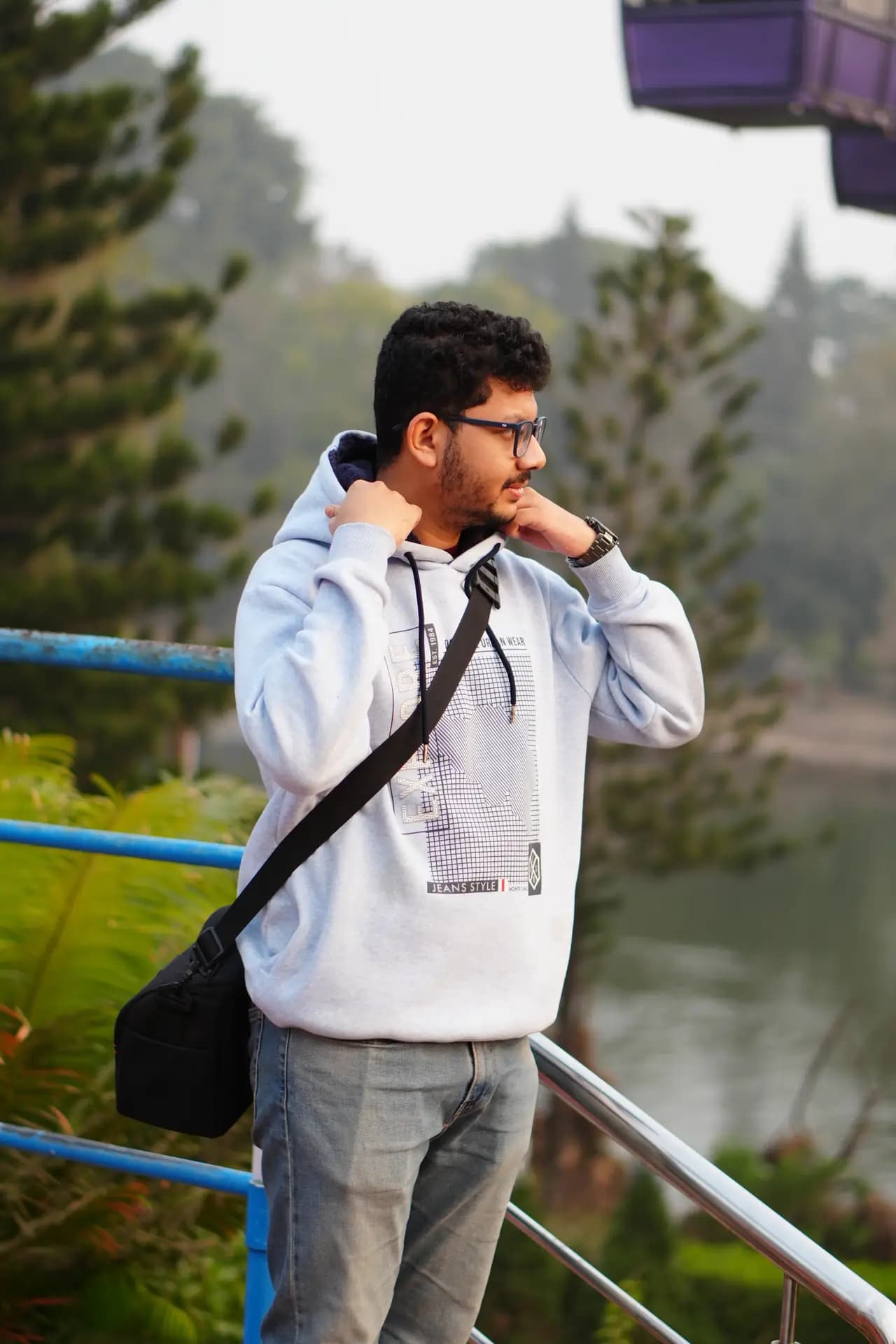 Raktim Ranjit in a hooded sweatshirt with a shoulder bag, standing by a blue railing above a lake