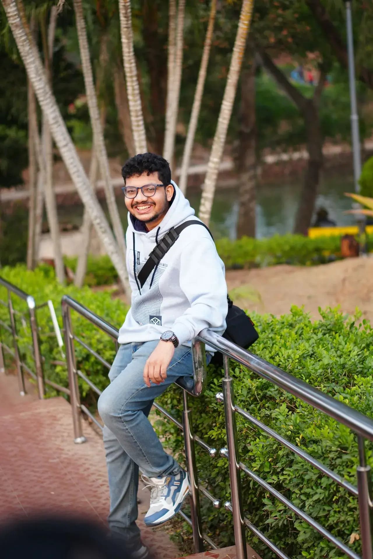 Raktim Ranjit smiling, leaning on a railing in a park, wearing a light hooded sweatshirt and a shoulder bag