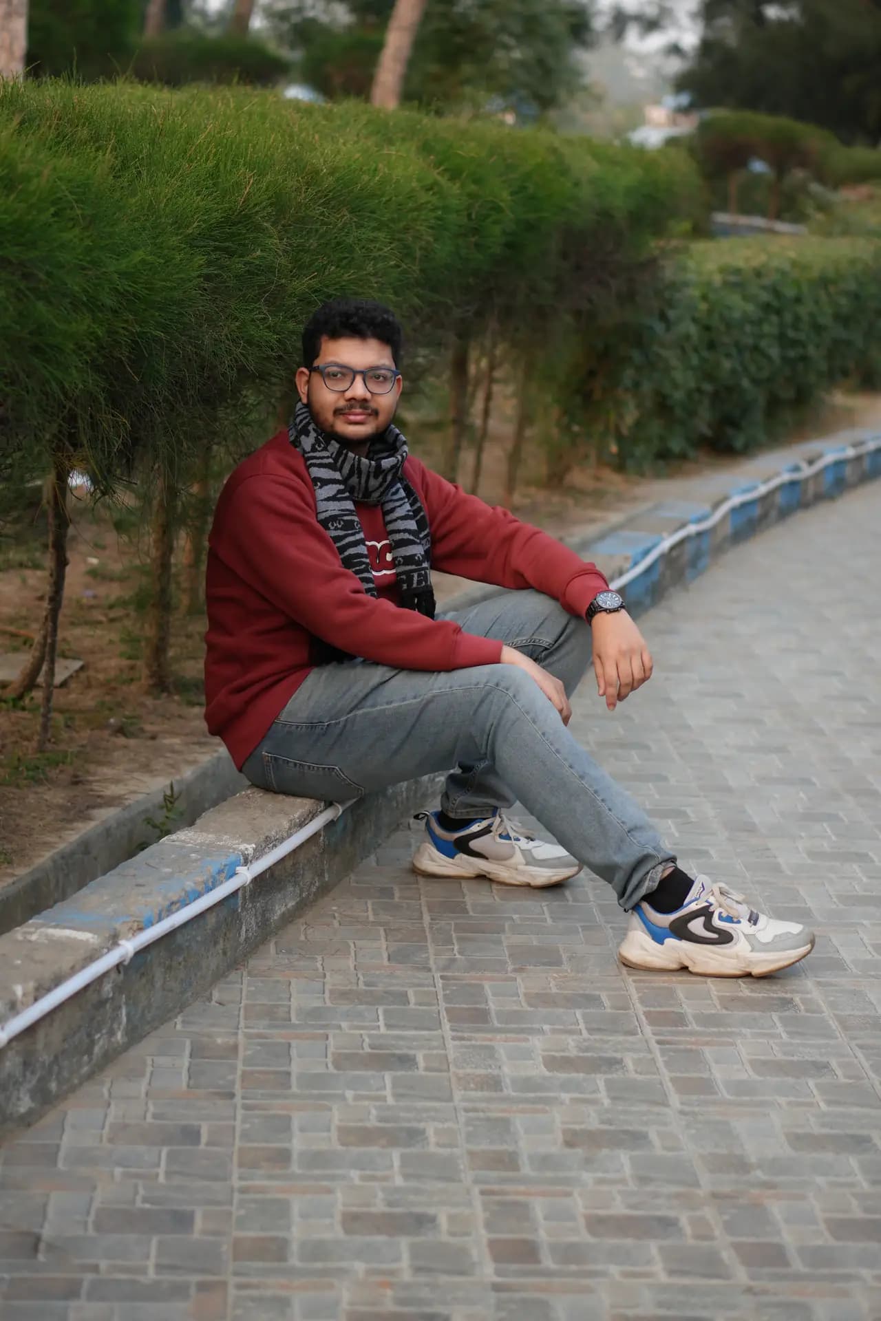Raktim Ranjit sitting on a low kerb beside a paved path lined with hedges